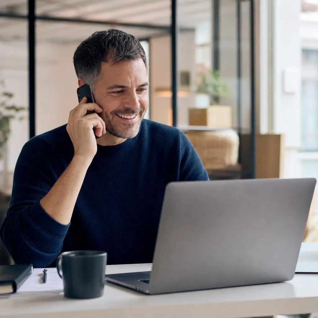 A relaxed business owner smiling on a phone call while looking at a laptop